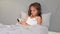 Smiling cheerful cute brown haired girl resting on pillow under blanket in her cozy bedroom using mobile phone watching cartoons