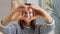 Smiling cheerful dark haired woman wearing striped shirt sitting indoor at home looking through heart shape making from hands