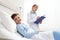 Smiling child lying in bed in hospital room looking at camera with doctor beside him filling out his medical record with copy spac