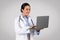 Smiling confident latin female doctor in white coat with stethoscope typing on laptop isolated on gray background