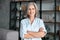 Smiling confident middle aged woman standing arms crossed in office, portrait.