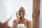 Smiling cute millennial african american lady with towel on head after shower applying cream on face