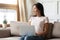 Smiling dreamy Asian young woman with laptop sitting on couch