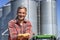 Smiling Farmer Showing Freshly Harvested Corn Maize Grains in front of Farm Grain Bins