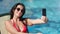 Smiling fashion woman in trendy sunglasses taking selfie using smartphone at sea water background
