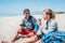 Smiling Father in sunglasses with smartphone sitting with teenager son with backpacks on the sandy seaside beach. Boy drinking wat