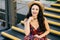 Smiling female with dark luxurious hair wearing dress and straw hat sitting at stairs looking with delightful look into distance.