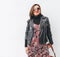 Smiling female dressed boho fashion style colorful long dress with black leather biker jacket with brown leather flap bag in