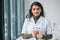 Smiling female physician posing in hospital office. Happy young indian woman doctor wearing white medical coat and