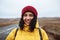 Smiling female tourist with a backpack wearing yellow jacket and red hat stands on the road. Young woman travels during winter or