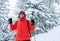 Smiling female trekker dressed red jacket in sunglases with trekking poles walking by snowy slope with fir-trees covered snow, Low