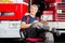 Smiling Firefighter Sitting On Chair Against Trucks