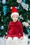 smiling funny Caucasian child girl toddler with blue eyes in red dress and Santa Claus hat sitting on gist box by New Year tree