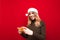 Smiling gamer girl in santa claus hat stands on red background with smartphone in hand, looks into camera and smiles. Happy lady