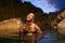 Smiling gender fluid black person poses in natural still water pool. Queer ethnic fashion model in open dress, brass