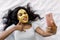 Smiling gesturing young African girl with golden facial mask, lies in white bed, smiling with phone in hands, while