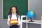 Smiling girl with backpack holding pile of books near blackboard at classroom. Cute pupil happy to go back to school