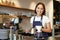 Smiling girl barista, asian bartender pouring water from kettle, brewing filter coffee in cafe behind counter, wearing