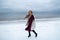 Smiling girl in the burgundy dress and coat on the background of the winter sea. Portrait of a woman on sea, snow windy