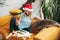 A smiling girl in a Christmas hat sits on the sofa and communicates via video link with friends, next to an open book and a gift.