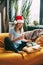 A smiling girl in a Christmas hat is sitting on the sofa and talking on the phone with friends, next to an open book and a gift.