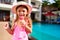 Smiling girl in pink swimsuit enjoys ice cream cone by poolside at summer resort. Child indulges in frozen treat on