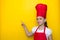 Smiling girl in a red chef`s suit points her finger at the space for lettering on a yellow background