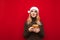 Smiling girl in santa claus hat stands on red background with smartphone in hand, looks away at blank space and smiles. Happy