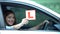 Smiling girl showing l-plate at car window, driving courses, left-hand traffic