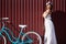 Smiling girl in white dress posing near blue bike in front of red fence.