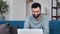 Smiling handsome Hispanic hipster man working chatting remotely use laptop sitting on blue couch