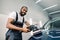 Smiling handsome young black skinned man, professional worker of car detailing service, polishing blue car with