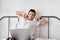 Smiling handsome young man in a white shirt and pajama sitting on a bed with laptop and stretching