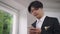 Smiling happy Asian groom in suit texting on smartphone standing indoors. Portrait of young man in wedding costume with
