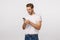 Smiling happy blond european guy in white t-shirt, jeans, holding smartphone, wear wireless headphones, look mobile