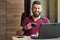 Smiling hipster keeping coffee cup, working with laptop in cafe.