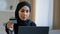 Smiling islamic girl customer in hijab sitting on sofa make online order use laptop enter credit card number for