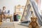 Smiling kid sitting on bench with notebook, real photo of natural playroom interior with scandinavian tent and teddy bear