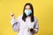 Smiling korean woman in medical mask, holding camera, asking to take photo, pointing at something on empty space, yellow