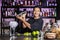 A smiling Latin American bartender mixes liquids with a cocktail shaker in front of a pub bar.