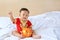 Smiling little Asian baby boy in traditional Chinese dress putting some coins into a piggy bank sitting on bed at home. Kid saving