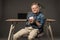 smiling little boy in eyeglasses sitting on chair with coffee cup near table with laptop and lamp on grey