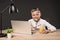 smiling little boy holding eyeglasses and sitting at table with glass of juice laptop plant and lamp on grey