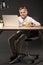 smiling little boy holding eyeglasses and sitting at table with laptop glass of juice plant and lamp on grey