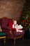 Smiling little boy reading book in front of christmas tree. Happy Small boy sits on checkered red armchair with book in living roo