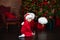 Smiling Little boy in red hat Santa sit on floor by Christmas tree with lantern. Happy small boy holding christmas lantern in livi