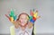 Smiling little cheerful girl showing her colorful hands and cheek painted in kids room. Focus at baby hands