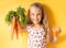 Smiling little girl in flowery dress showing to camera a bunch of delicious carrots and glass of carrot juice. Waist up