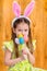 Smiling little girl with long blond hair wearing pink and white rabbit or bunny ears and holding bunch of painted colorful eggs