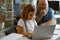 Smiling little girl types on laptop while father sits nearby at table in light room